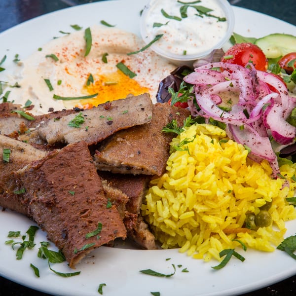 A gyro platter with sliced meat, yellow rice, salad, hummus, and tzatziki sauce.