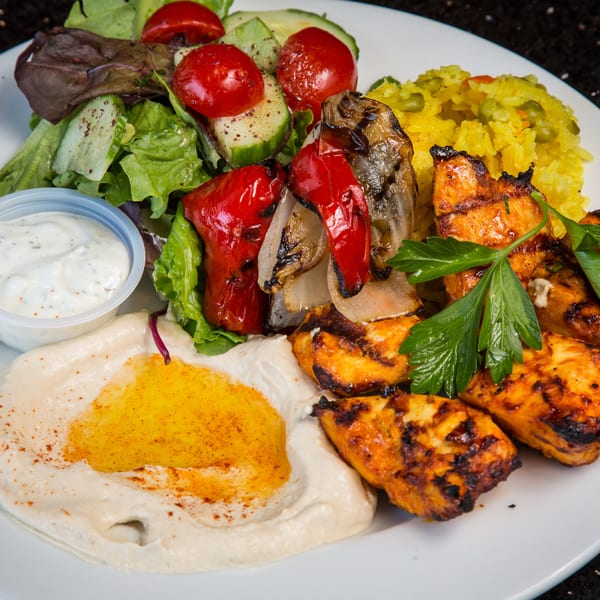 A plate of grilled chicken, hummus, rice, salad, and roasted vegetables with a side of sauce.